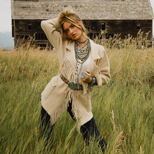 A woman posing in a tall grassy field with a rustic wooden barn in the background, wearing a cream-colored fringed cardigan, layered turquoise and silver necklaces, bracelets, and a belt with a turquoise buckle, paired with dark pants.