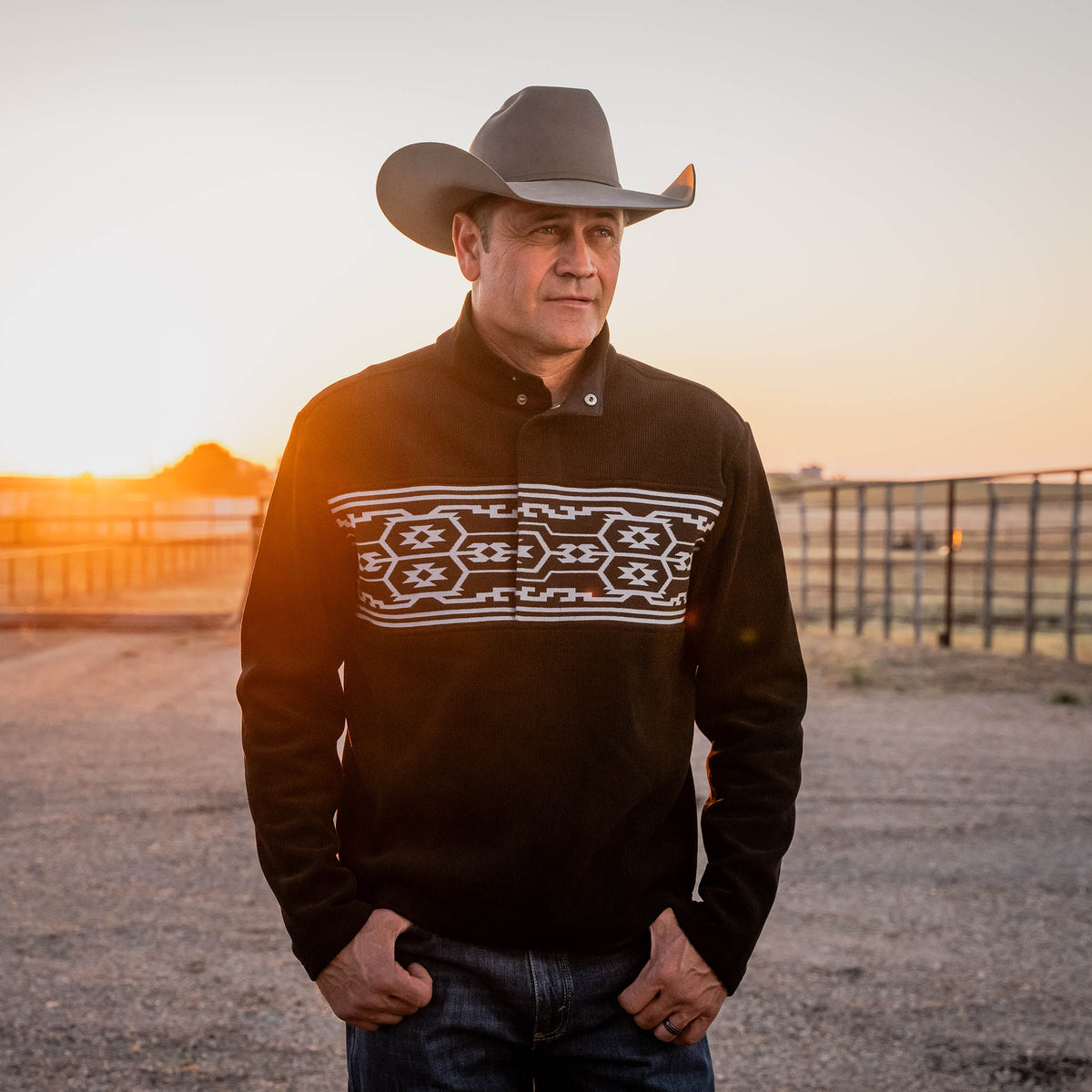 Man wearing a cowboy hat and patterned sweater standing in an open field with sunset.