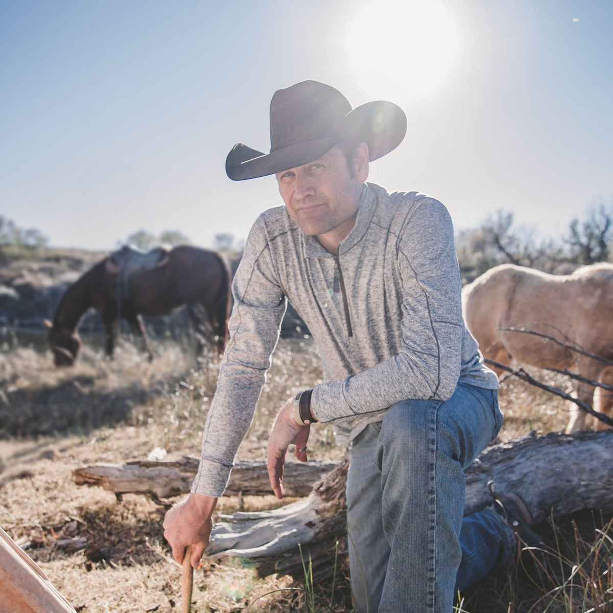 Stran Smith kneeling on the ground in front of horses wearing a felt cowboy hat and a gray quarter zip lightweight jacket.