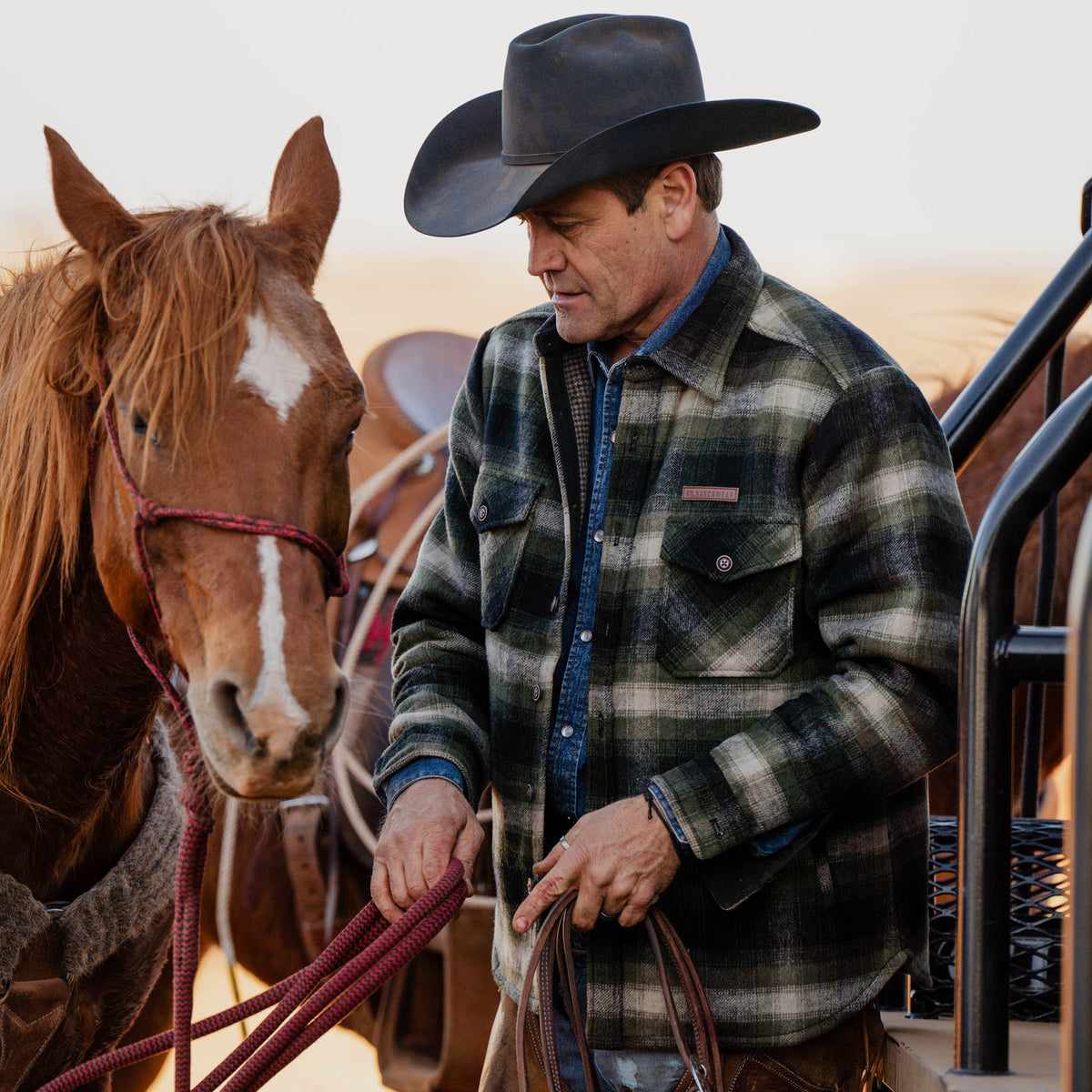 Man in a plaid shirt and cowboy hat standing next to a horse, holding reins.