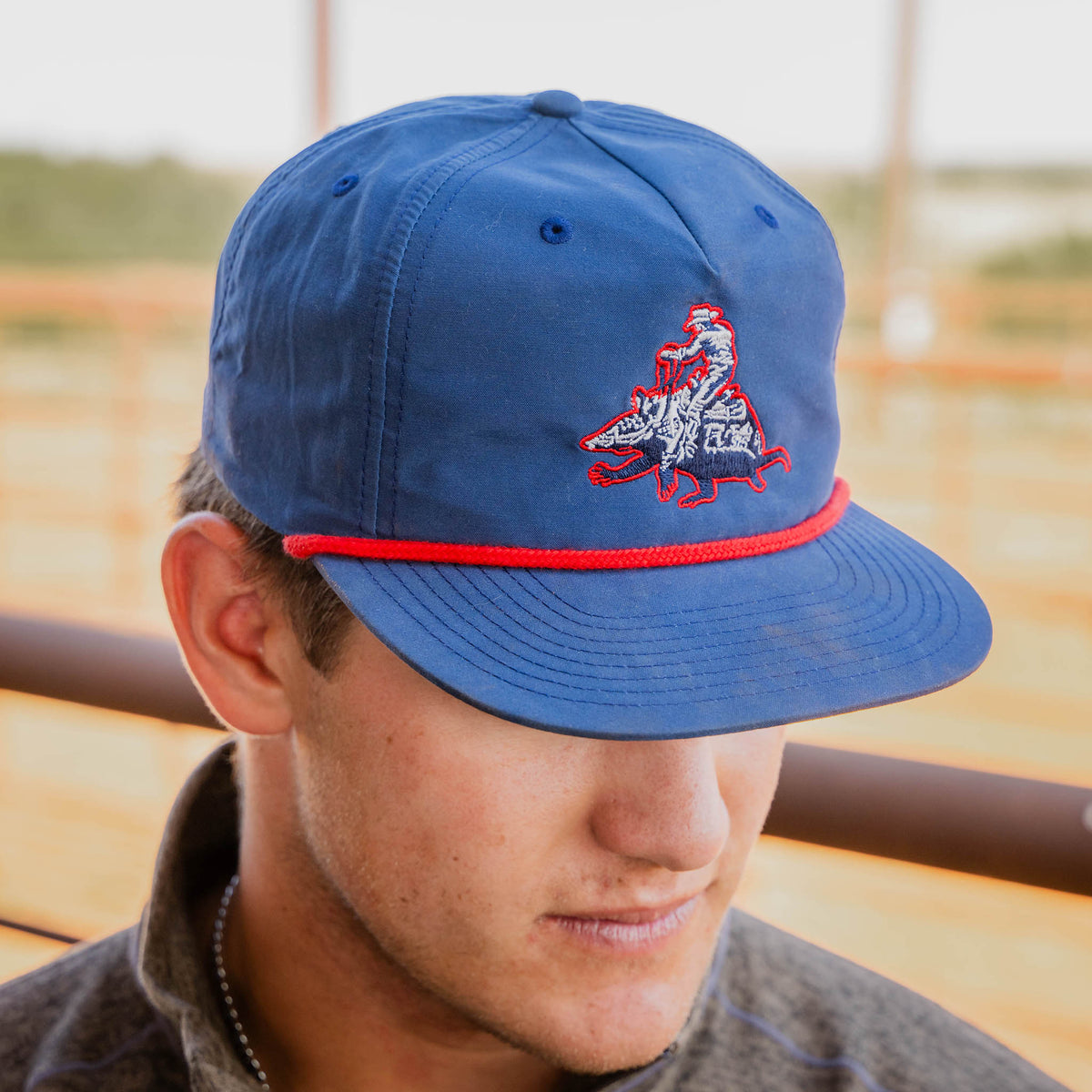 Man wearing a flat bill navy rope hat with red braided rope detail and STS Ranchwear embroidery on the front with a blurred background.