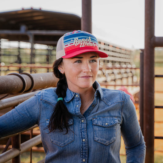 Woman wearing a denim long sleeve shirt and a trucker style hat with geometric pattern featuring stripes and geometric shapes in multiple colors, including red, blue, beige, and green, with 'Ranch Wear' text embroidered in the center.