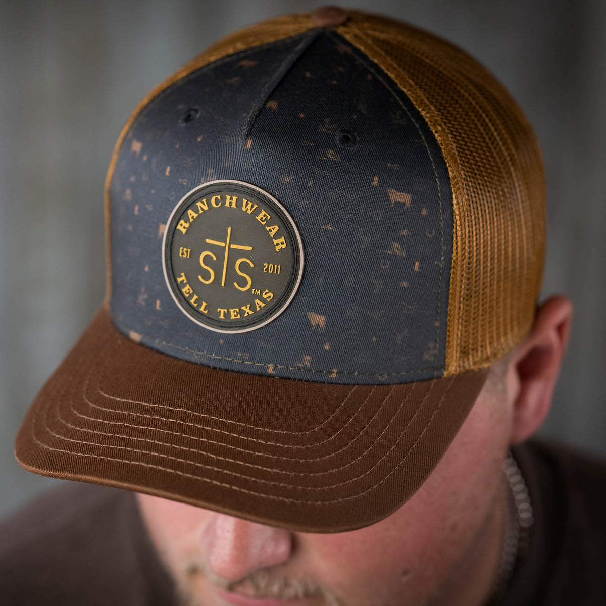 A black and brown trucker style hat with an STS Ranchwear rubber patch and an adjustable plastic snapback.