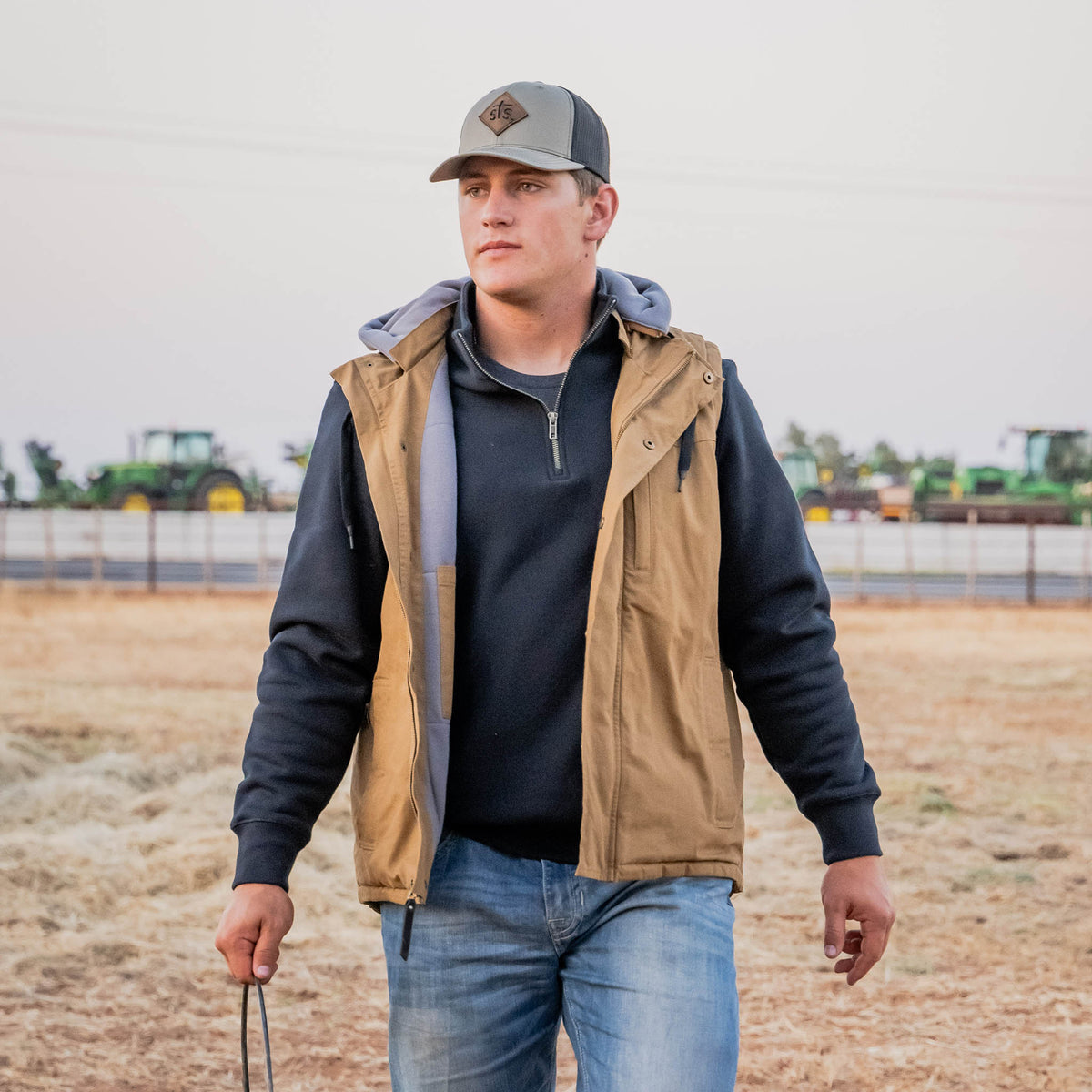 Man wearing a brown vest and cap standing in an open field with tractors in the background.