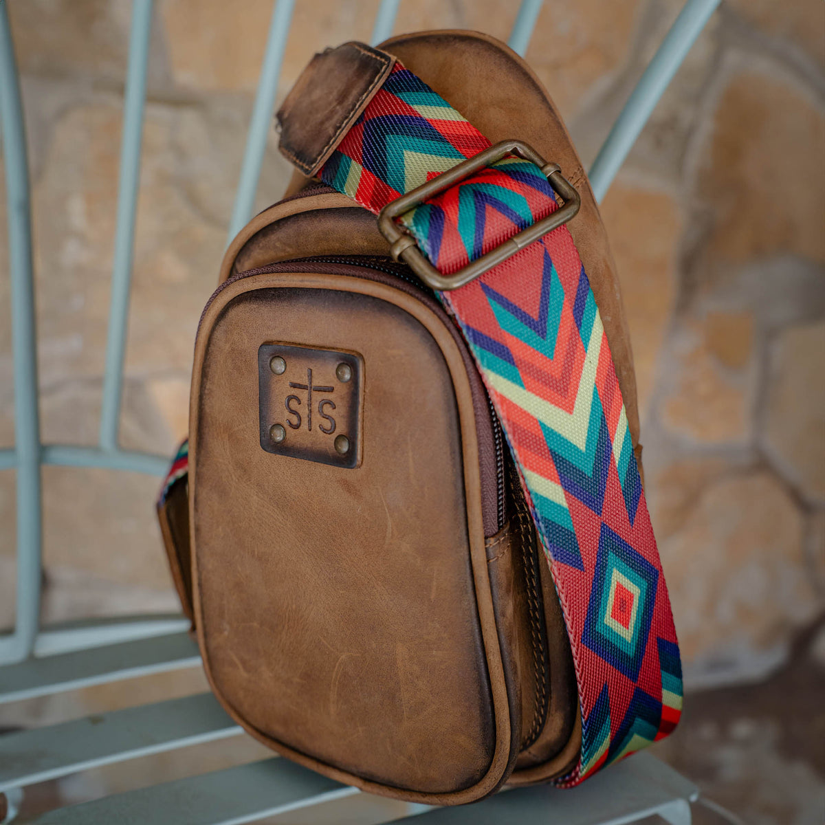 Brown leather sling bag with colorful nylon strap on a chair.