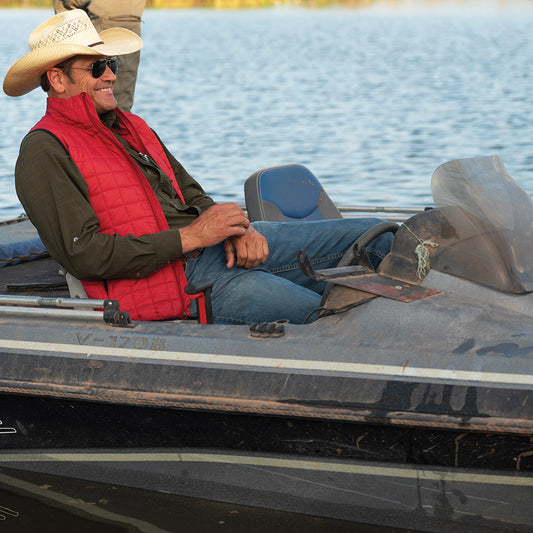 Man wearing a red quilted lightweight jacket, blue jeans and tan cowboy hat sitting on a boat.