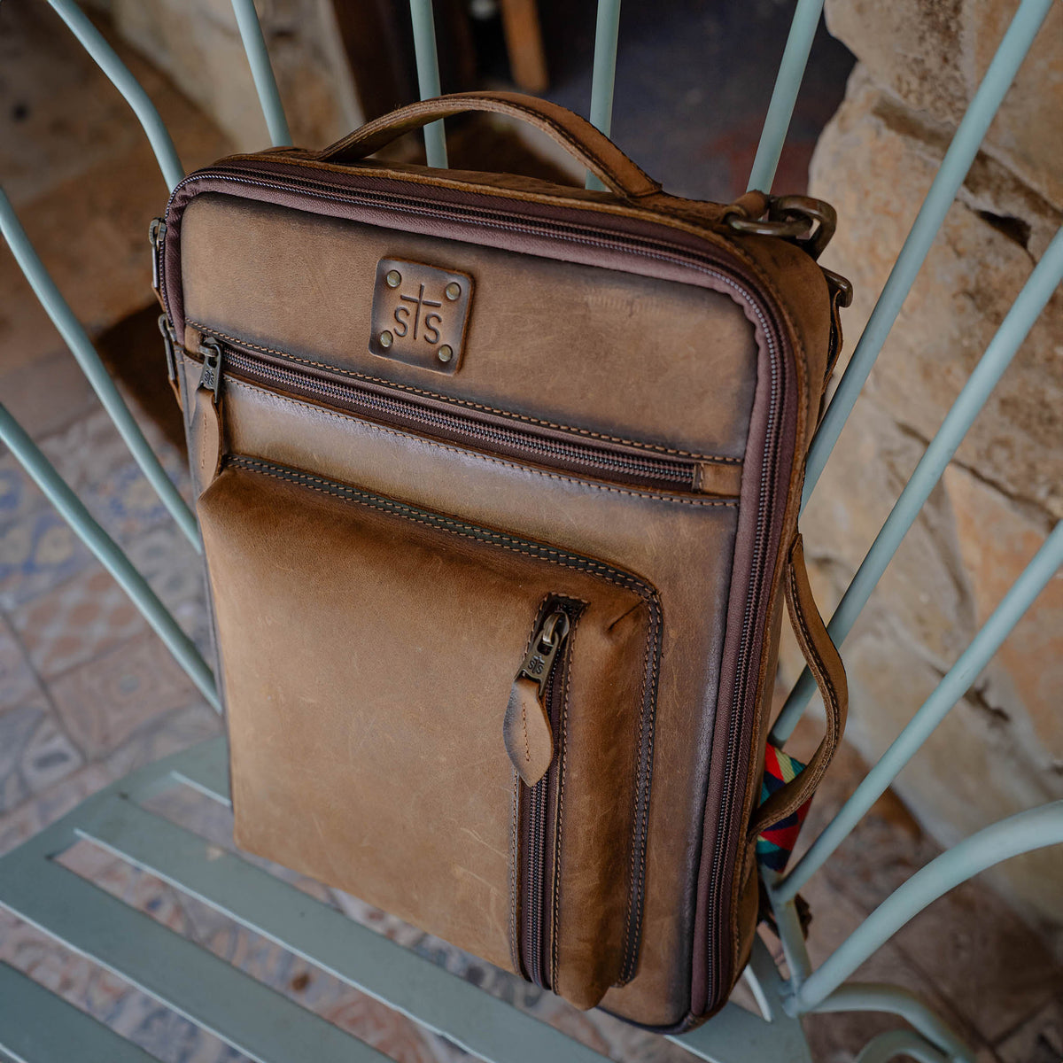Brown leather messenger bag with multiple zip pockets and leather handle sitting on a metal chair.