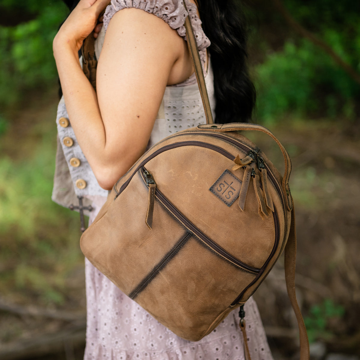 Person standing in the woods with a brown genuine leather backpack over their shoulder.