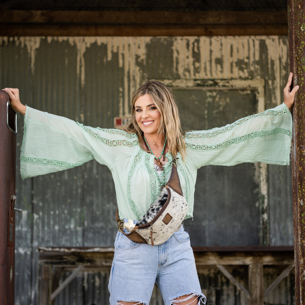 Woman wearing green shirt and jeans leaning between two poles with a cowhide belt bag across her body.