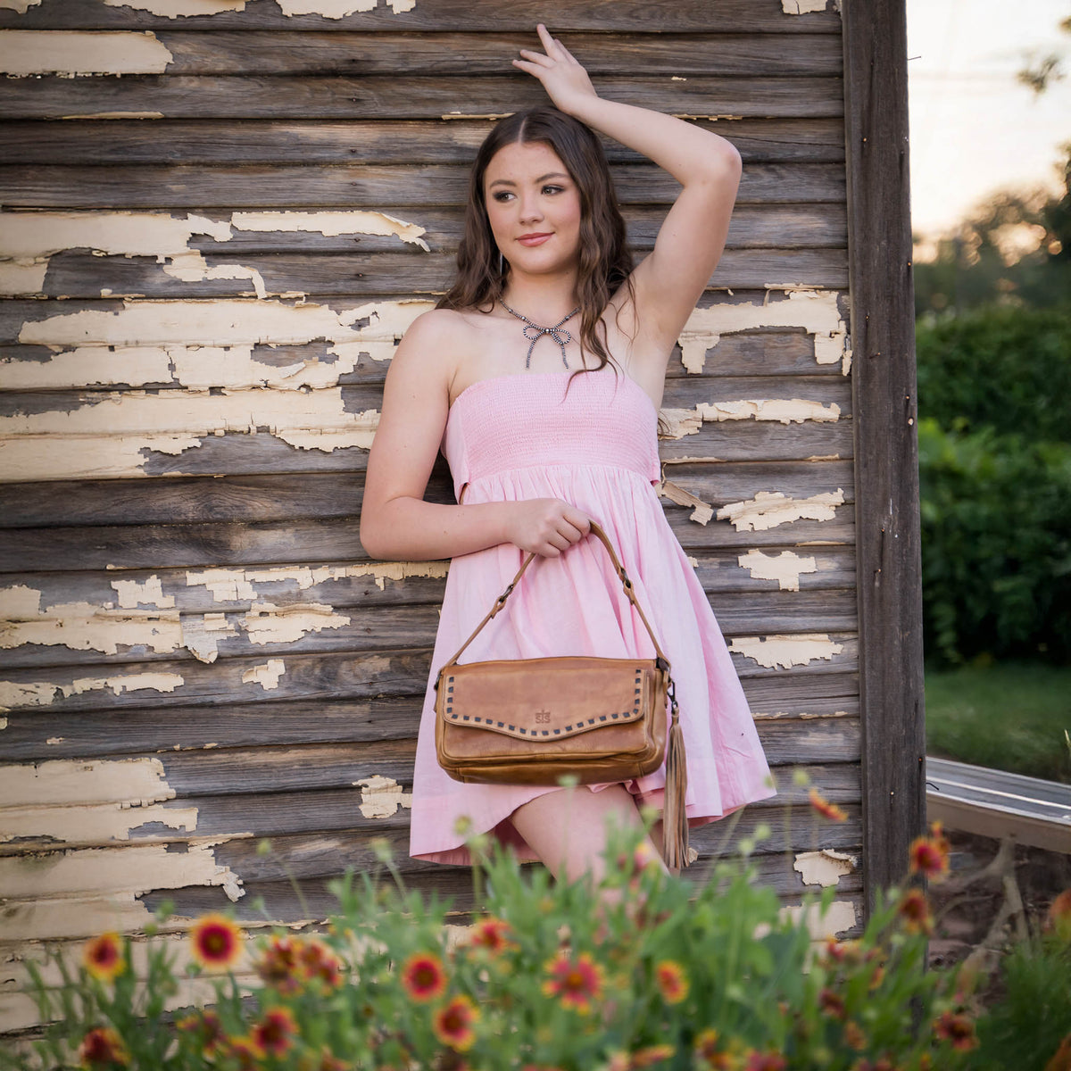 Woman in a pink dress holding a brown leather handbag against a wooden wall with plants in the foreground.