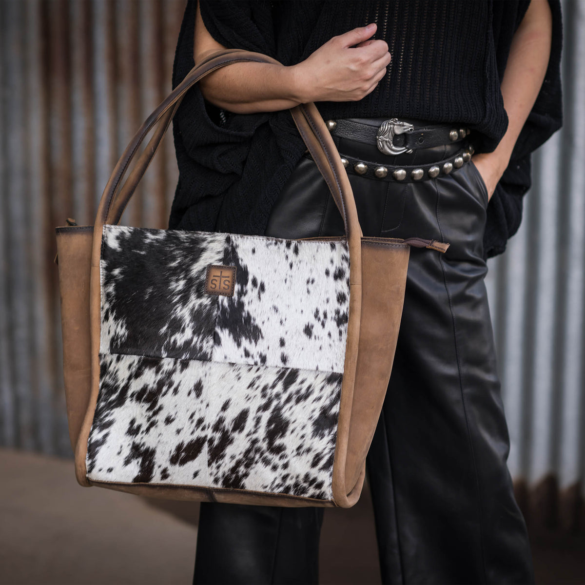 Person holding a genuine leather tote bag with black and white hair on hide cowhide accents and dual rolled leather handles.