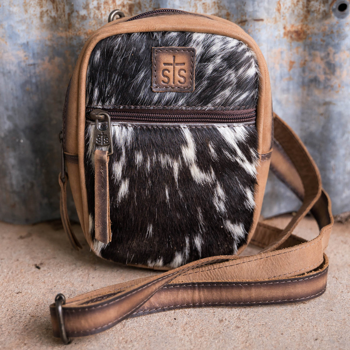 A cowhide crossbody bag with full grain leather accents, black and white hair on hide cowhide, featuring a zip closure, a front exterior zip pocket and a brown leather strap.