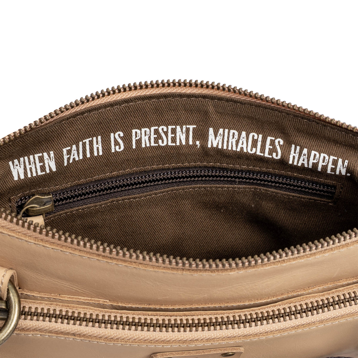 A beige leather crossbody with the quote: "when faith is present, miracles happen." in the lining of the top zip closure on a white background.