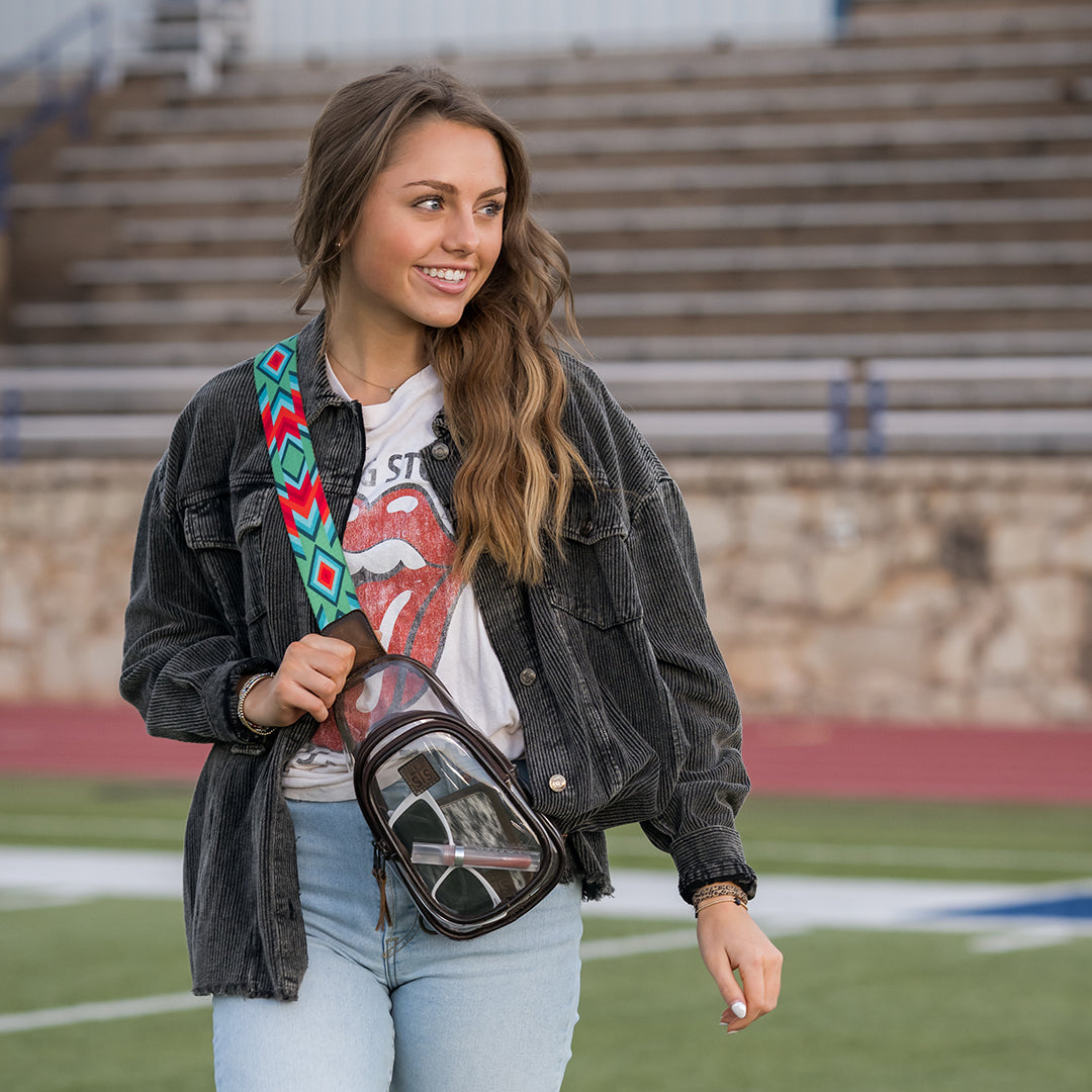 Woman holding a transparent sling bag with a colorful strap on a sports field.
