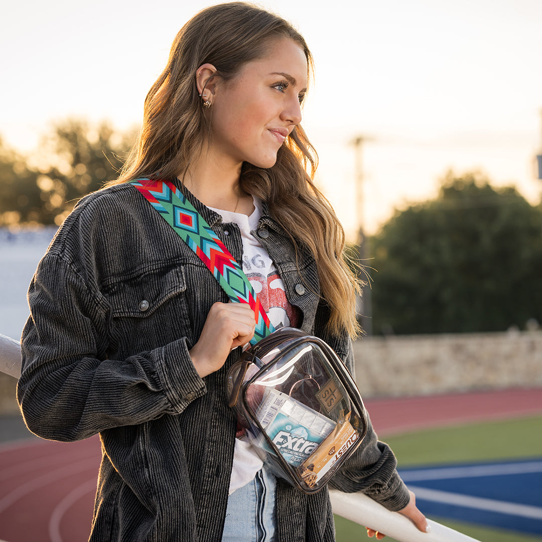 Woman holding a clear bag with a visible brand logo and colorful nylon strap on a sports field.