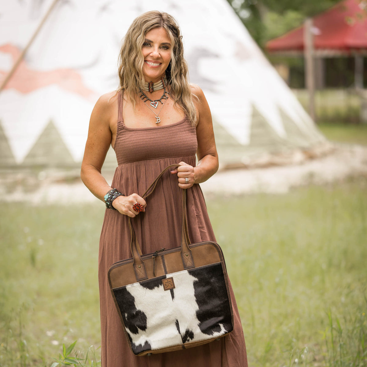 Woman holding a brown leather shopper bag with black and white cowhide accents in a natural outdoor setting.