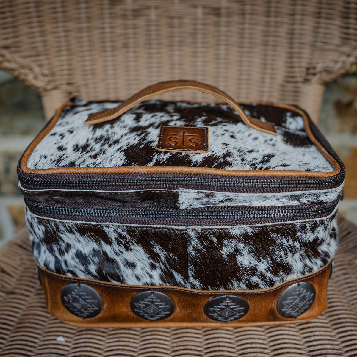A cowhide makeup organizer with brown leather accents and large rustic conchos, featuring a zip around top closure and a leather handle.