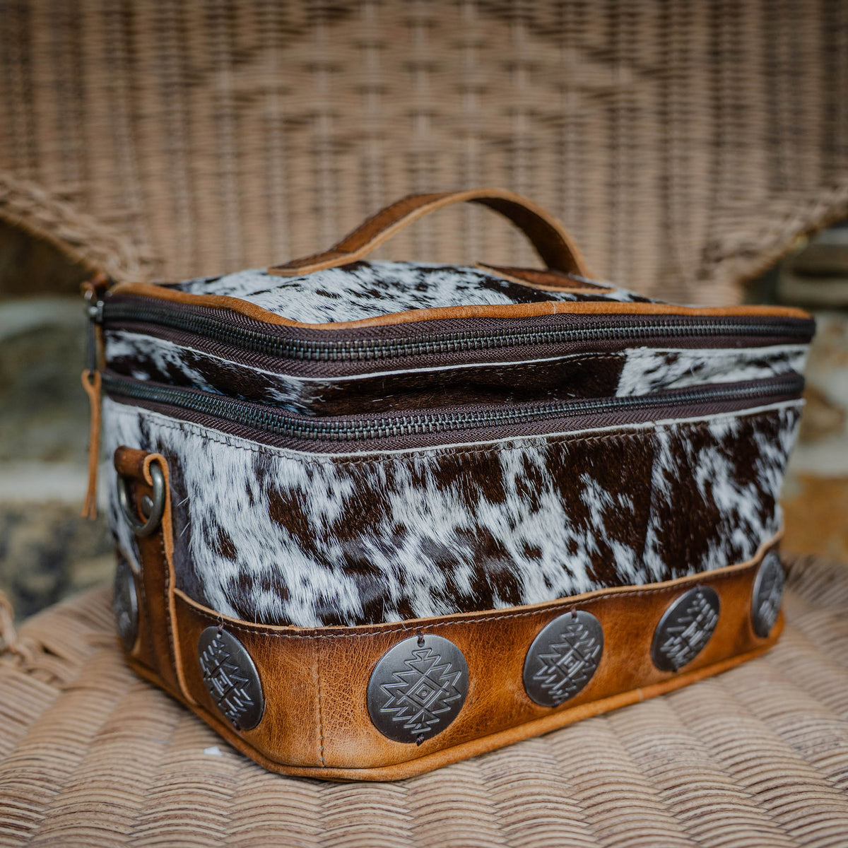 A cowhide makeup organizer with brown leather accents and large rustic conchos, featuring a zip around top closure.