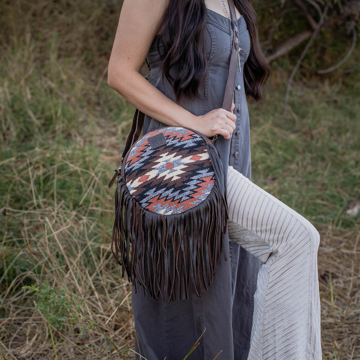 Person holding a round, aztec patterned bag with leather accents and fringes outdoors.