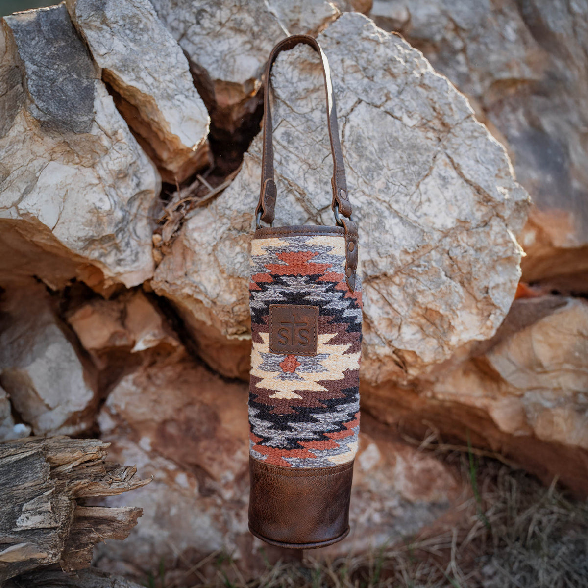 Patterned wine bottle holder with leather strap on a rocky background.