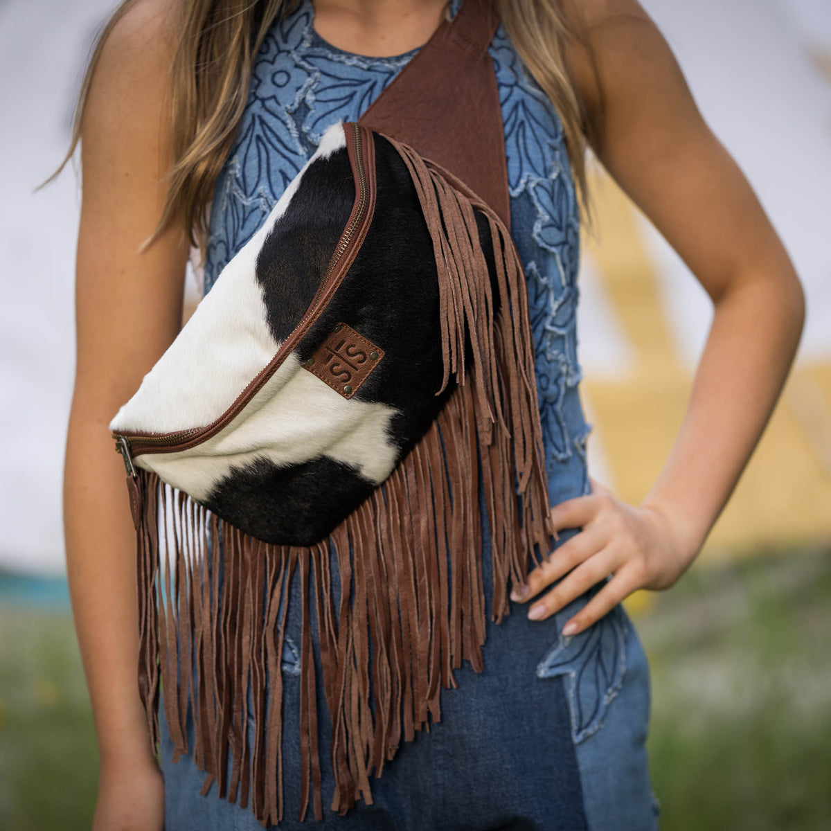 Person wearing a cowhide leather belt bag with fringe and a brown leather patch with embossed STS Ranchwear logo.
