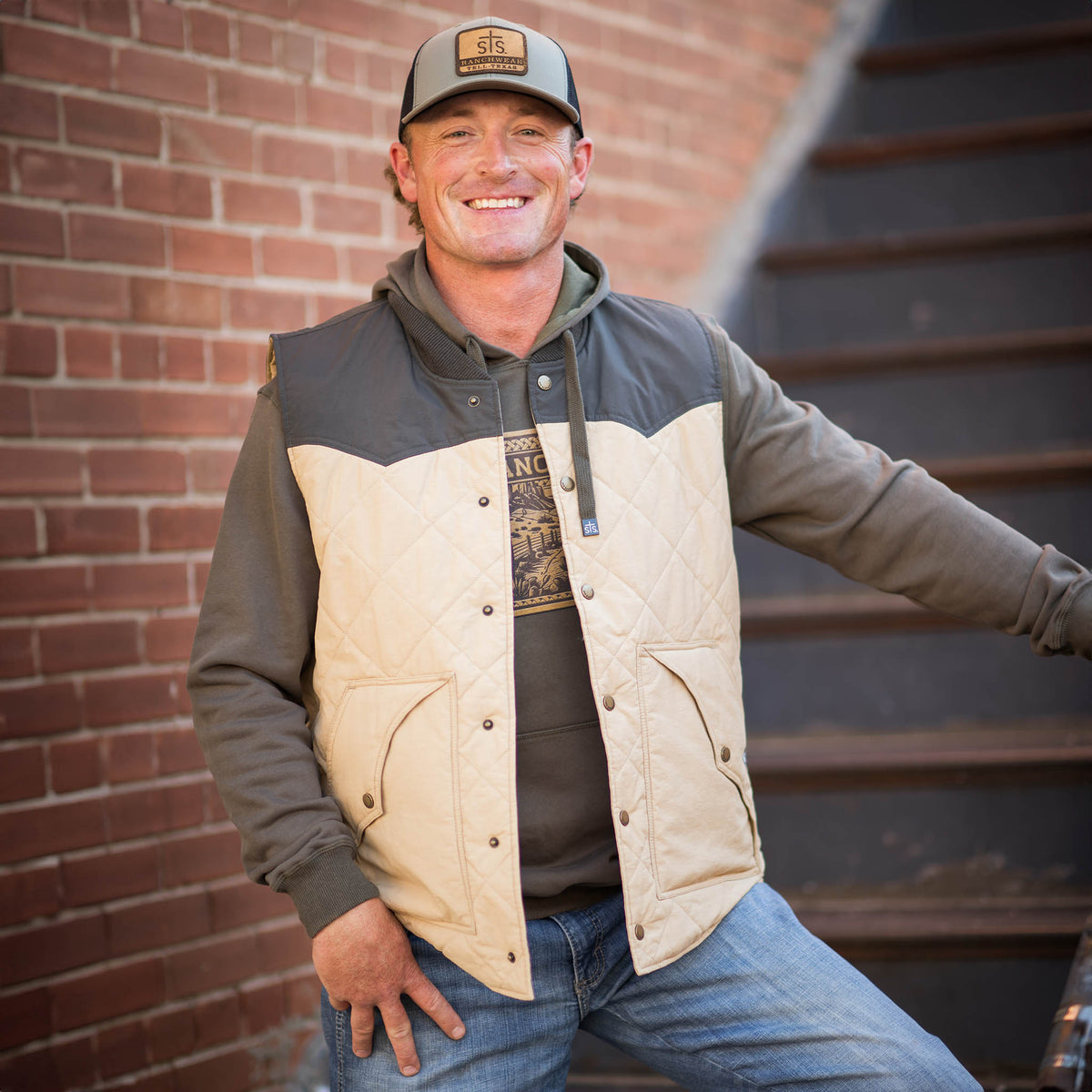 Man standing on stairs wearing a men's Pagosa Vest featuring a quilted design, snap front closure, and two exterior hand warming pockets. The vest has a beige shell with a dark gray western yoke.