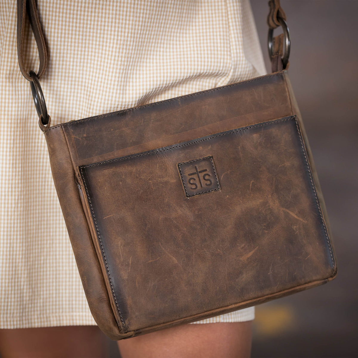 A leather purse with top zip closure, magnetic front pocket and adjustable crossbody strap rested against a woman's plaid yellow dress.