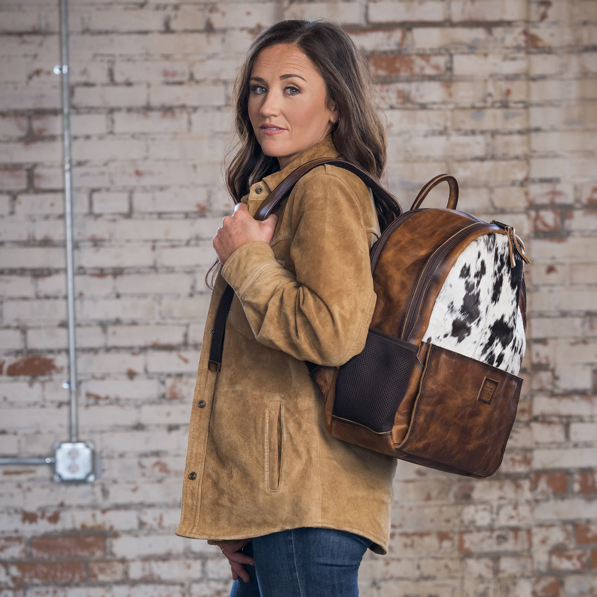 Yaleigh Yarbrough wearing our distressed full grain leather and cowhide backpack with double zip around closure, top carry handle and adjustable straps in front of a brick wall