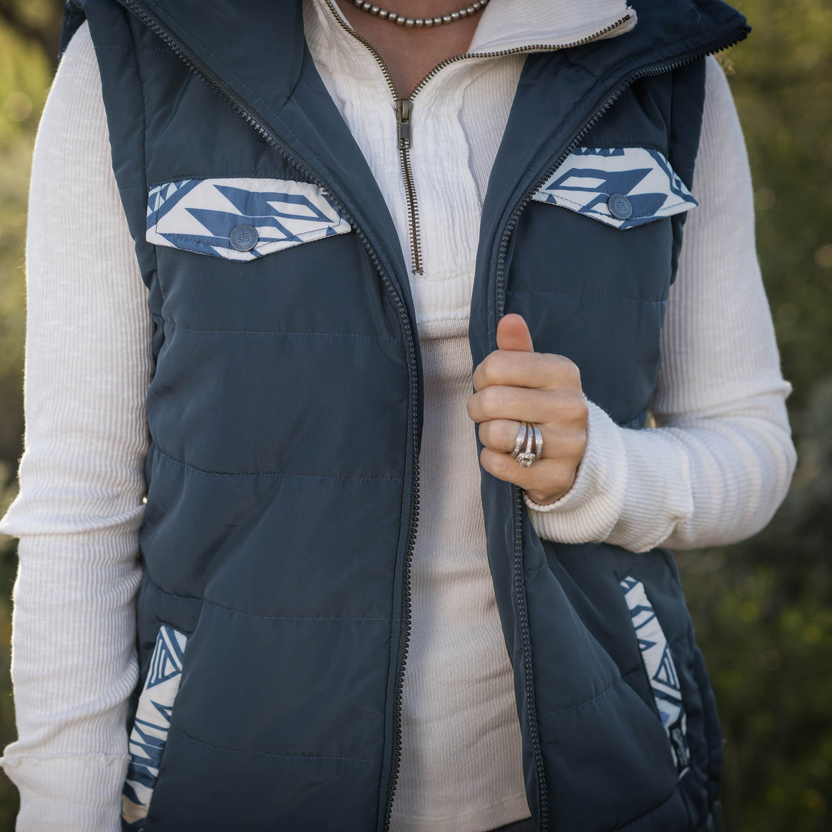 Person in a forest setting wearing a white long sleeve shirt and navy puffer style women's vest with zip front closure and decorative aztec accents on chest and hand pockets.