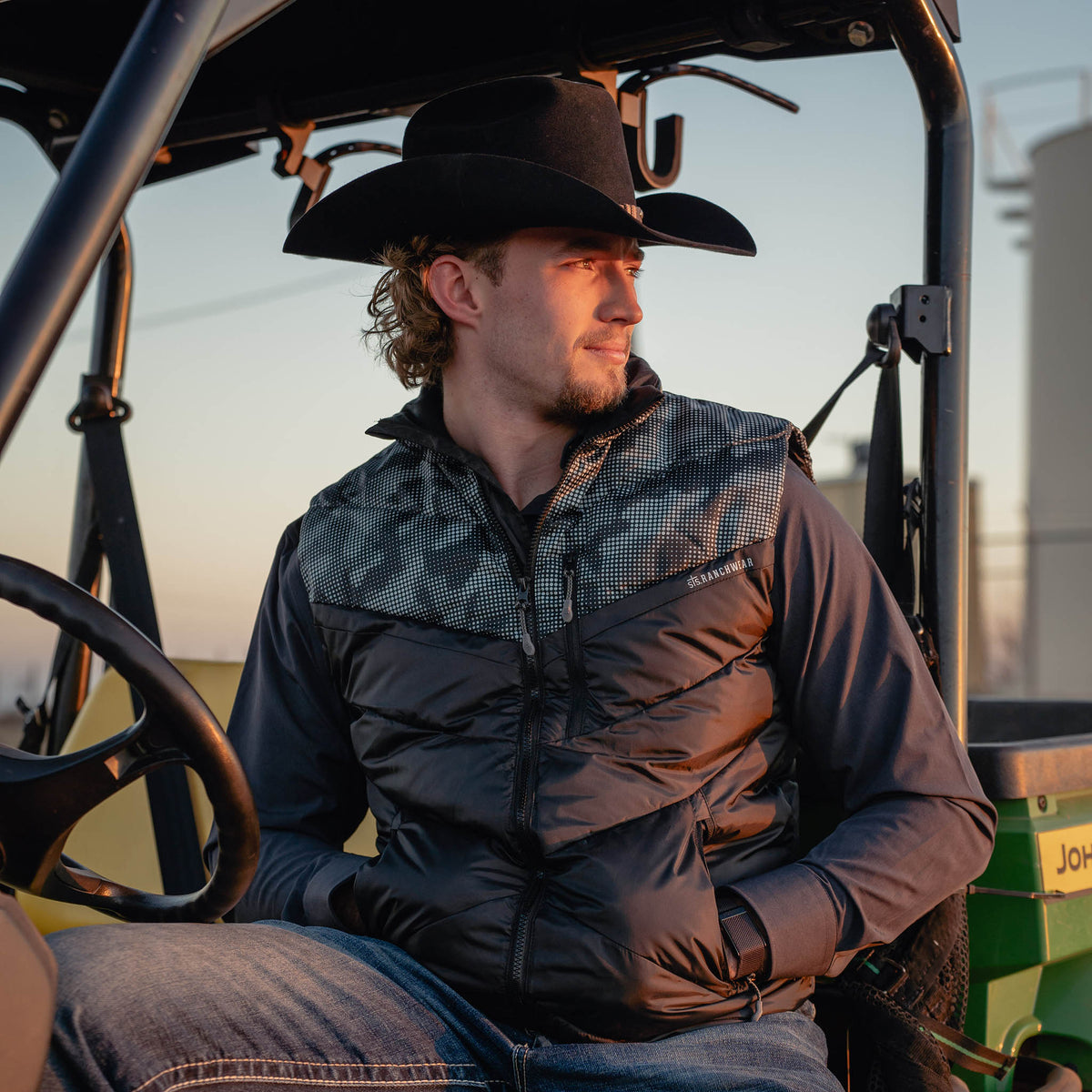 Man wearing a cowboy hat and puffy vest sitting inside a vehicle with a sunset in the background.