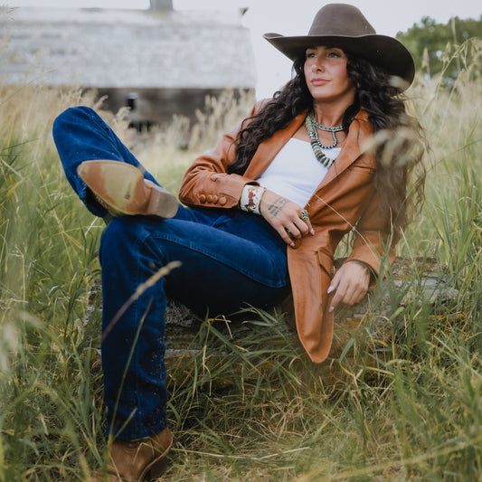A woman sits in tall grass outdoors wearing a brown wide-brim hat, tan leather jacket, blue jeans, white top, and brown boots, with layered necklaces and bracelets.