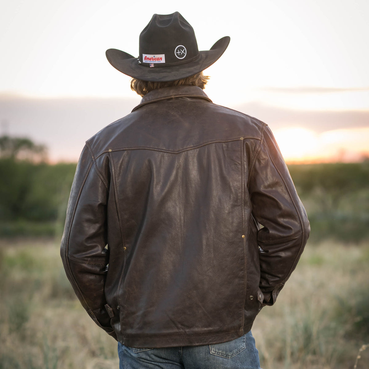 Man facing away standing in a field wearing a black cowboy hat, jeans and brown genuine leather Rifleman jacket.