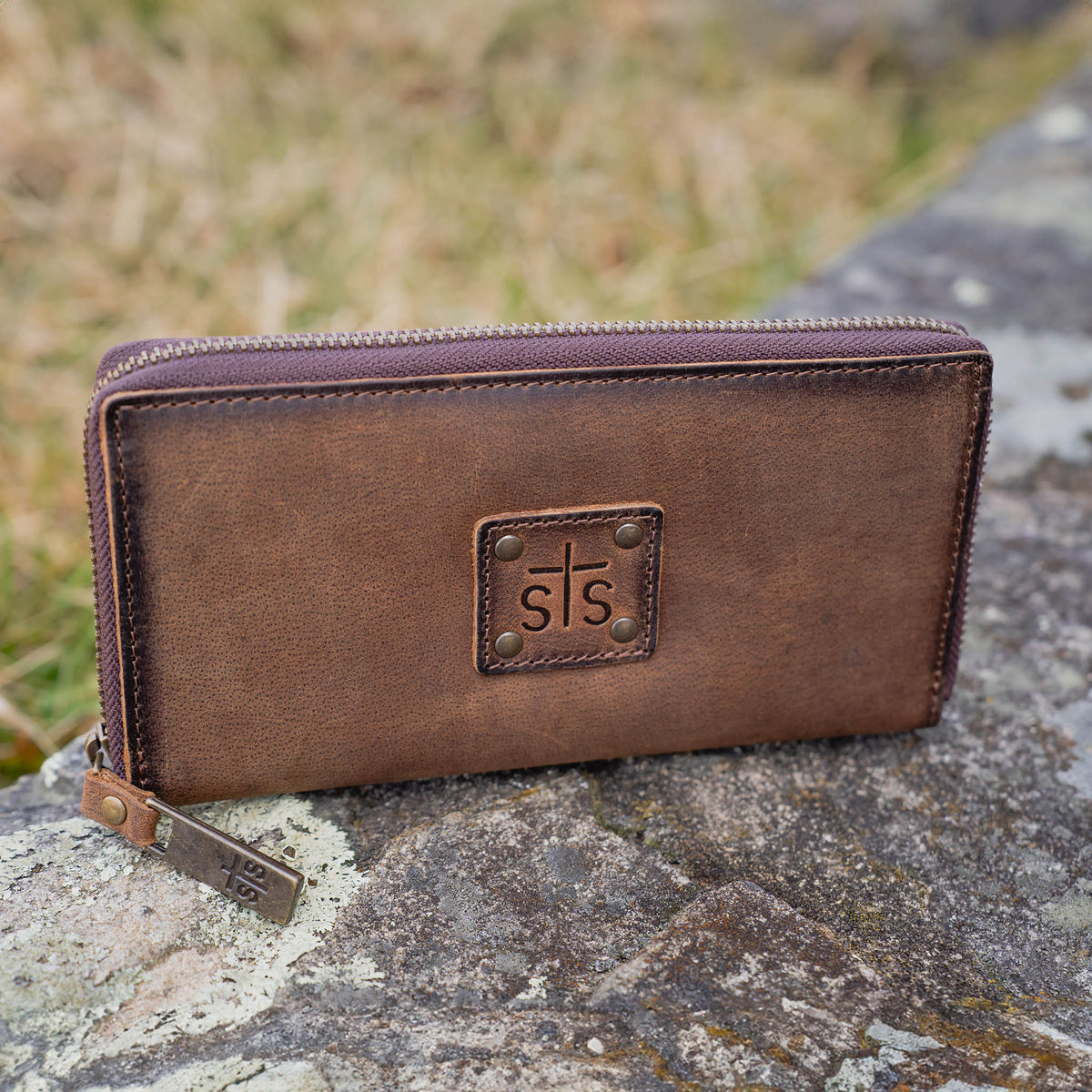 Brown leather wallet with zip around closure and "STS" stamped on a leather patch sitting on top of a rock.