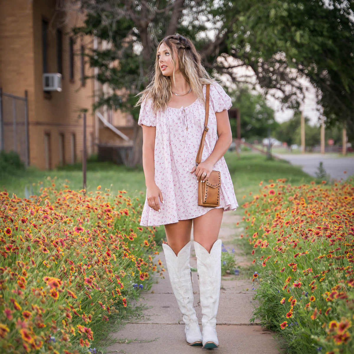 A woman wearing a Bandera tan leather phone sling wallet with buckstitch, zip around closure, magnetic flap for phone, and adjustable crossbody strap on the sidewalk next to a patch of flowers. She has on tall white cowgirl boots and a pink floral baby doll dress.