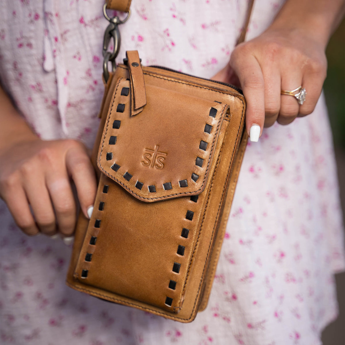 Closeup view of a woman's manicured hands holding a Bandera tan leather phone sling wallet with buckstitch, zip around closure, magnetic flap for phone, and adjustable crossbody strap. She is wearing a pink floral baby doll dress.