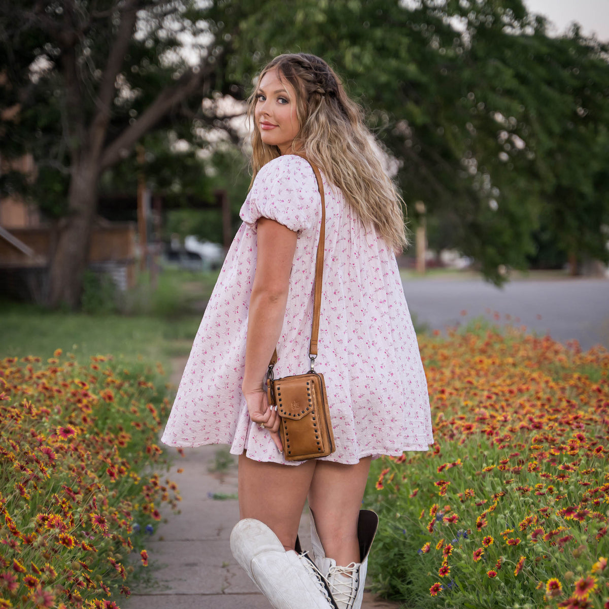 A woman wearing a Bandera tan leather phone sling wallet with buckstitch, zip around closure, magnetic flap for phone, and adjustable crossbody strap on the sidewalk next to a patch of flowers. She has on tall white cowgirl boots and a pink floral baby doll dress.