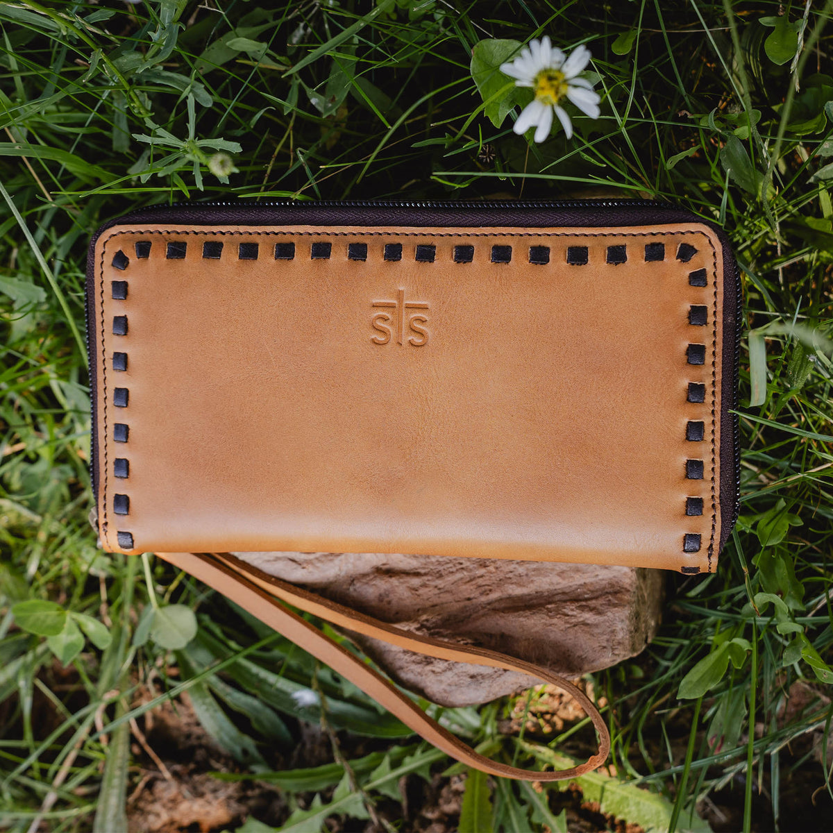 Bandera tan full grain leather wallet with buckstitch, zip around closure and wristlet strap on a rock in a patch of grass.