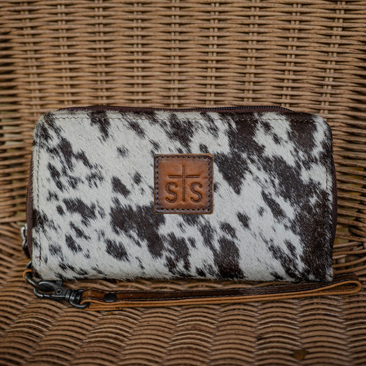 A cowhide wallet with a brown and white pattern, featuring a leather wristlet strap and the 'STS' brand logo embossed on a leather patch.