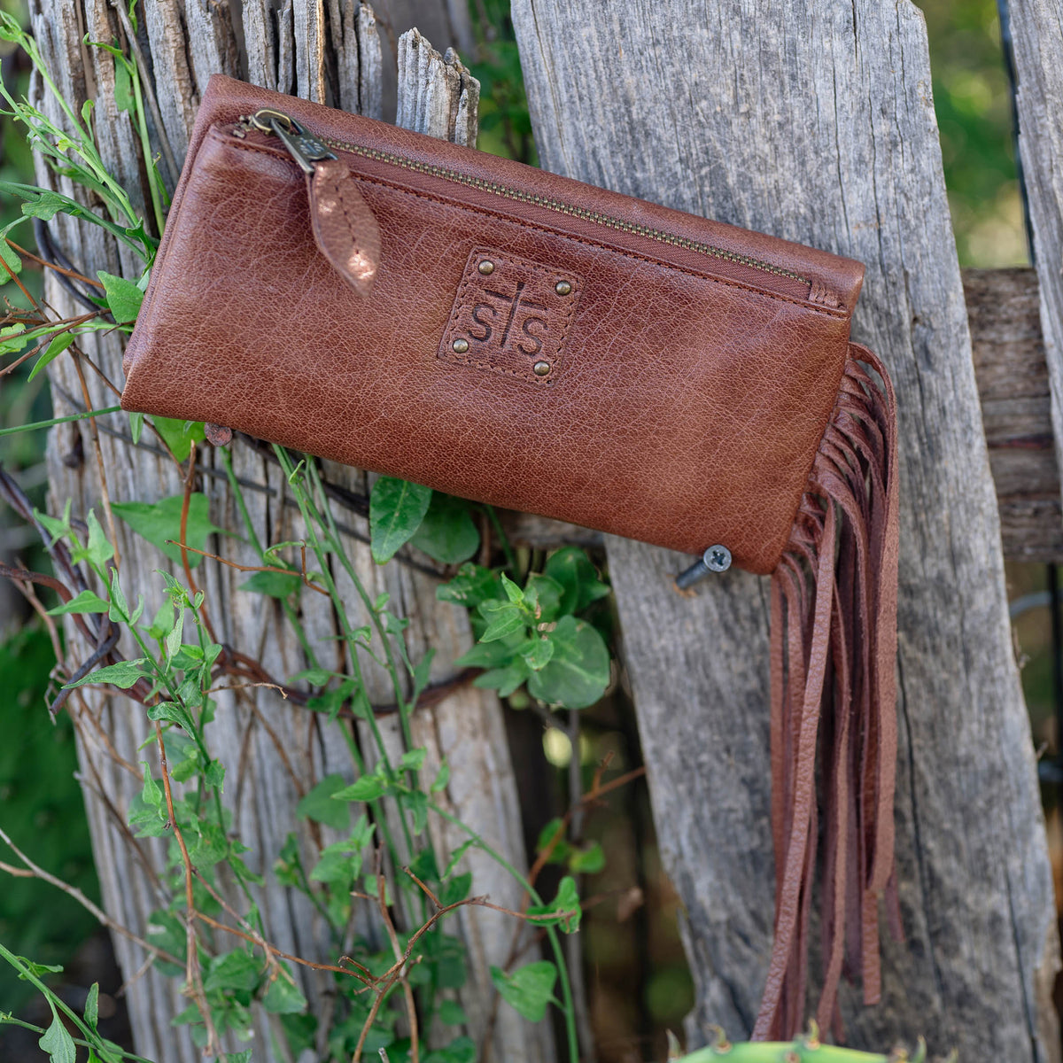 A brown buffalo leather wallet with fringe accents, featuring a fold and magnetic snap closure and exterior zip pocket sitting on a wooden fence post.