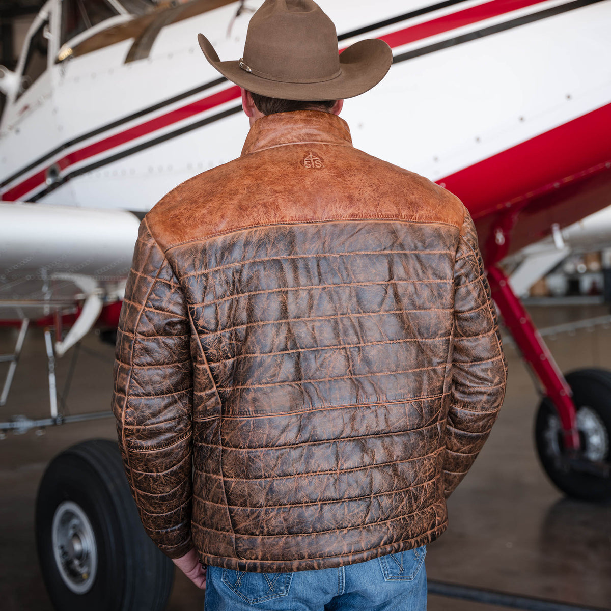 Stran T Smith facing away wearing a tan cowboy hat, jeans and a brown men's Clyde leather puffer jacket with a full-grain leather exterior standing in front of a small airplane.