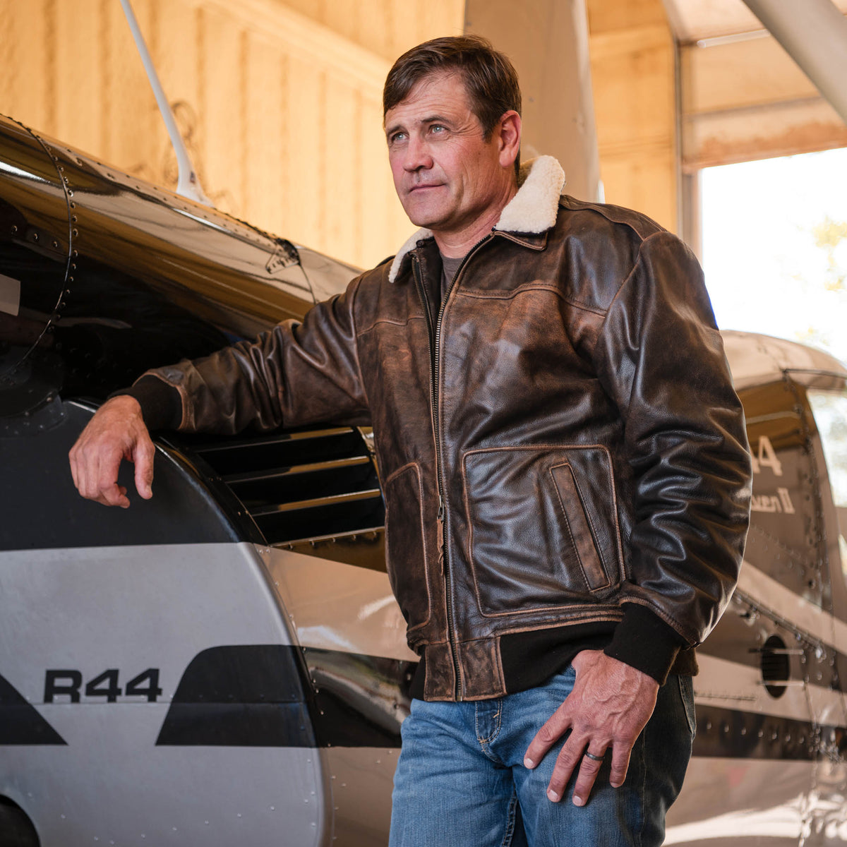 Stran T Smith leaning against a small airplane wearing blue jeans and a brown genuine leather bomber jacket with a removable sherpa lined collar and front zip closure.