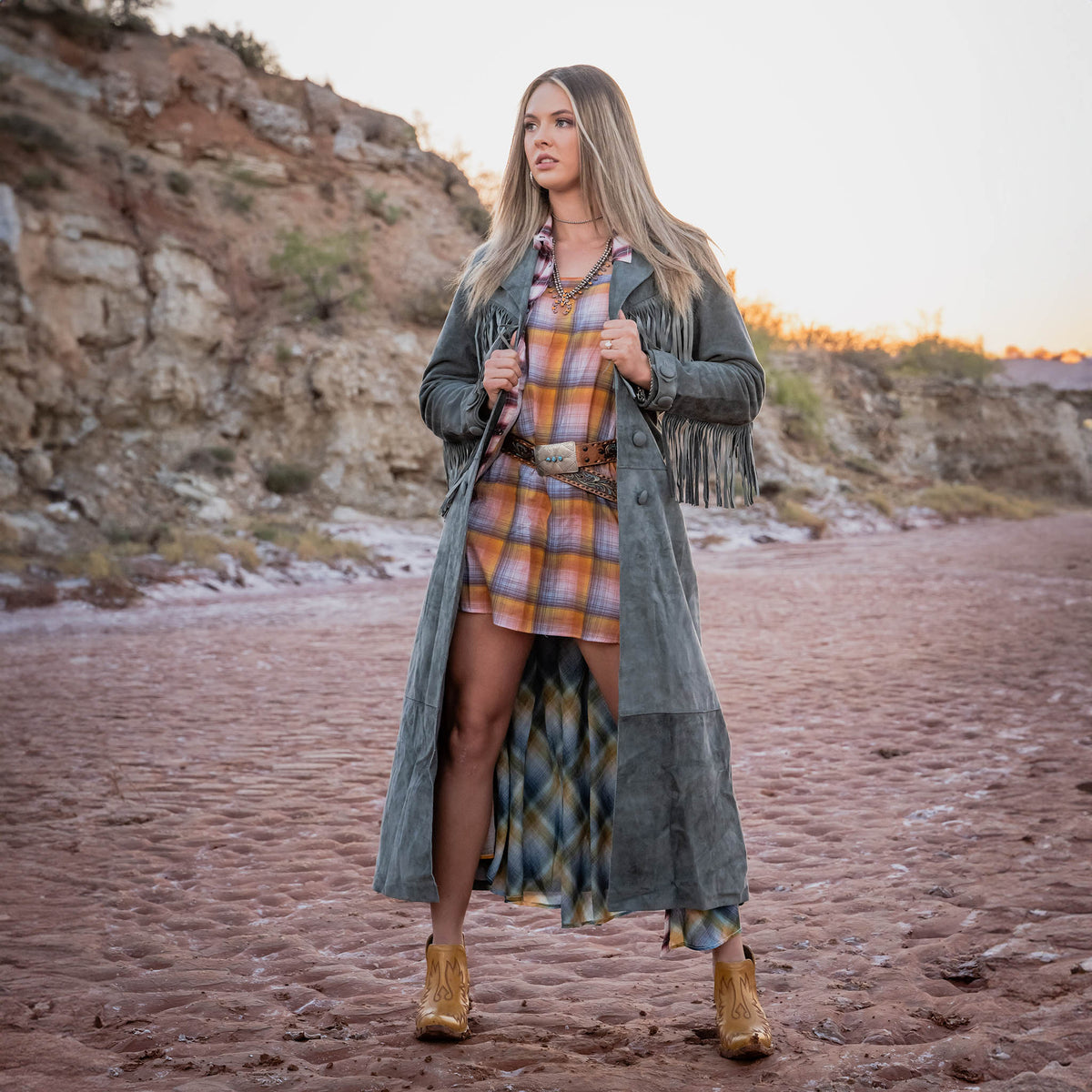 Woman in a plaid dress and grey leather suede trench coat with fringe standing in a desert landscape.