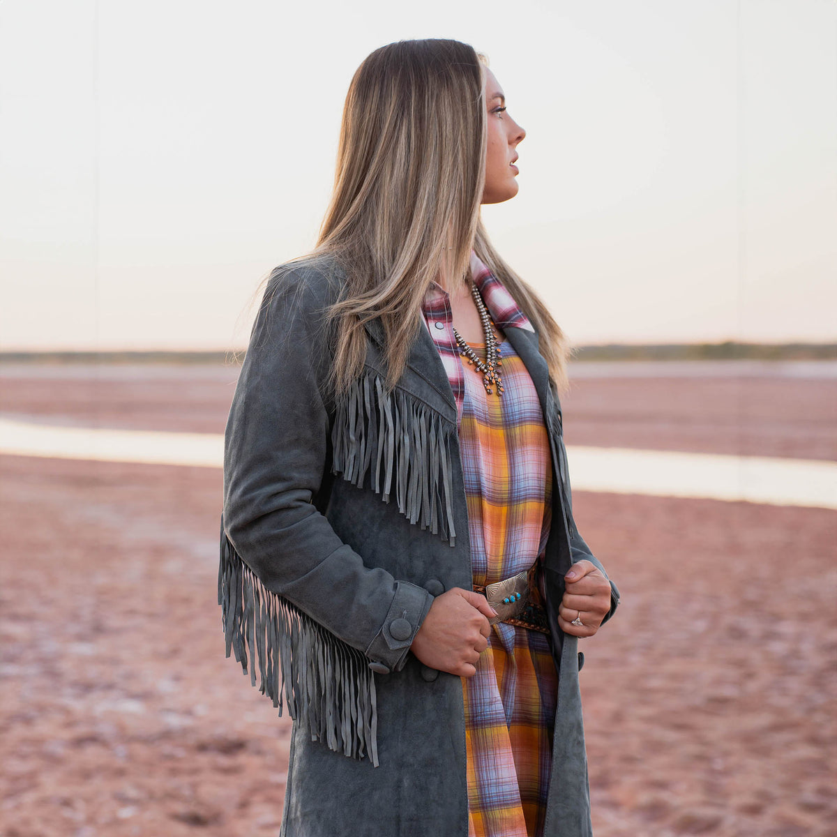 Woman wearing a gray fringed suede leather jacket and plaid dress standing on a flat, open landscape.