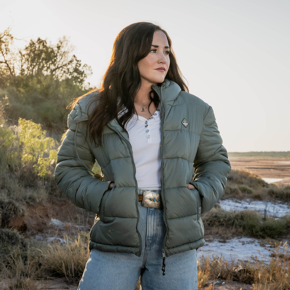 Woman wearing a green puffer jacket standing in a natural setting with trees and open space.