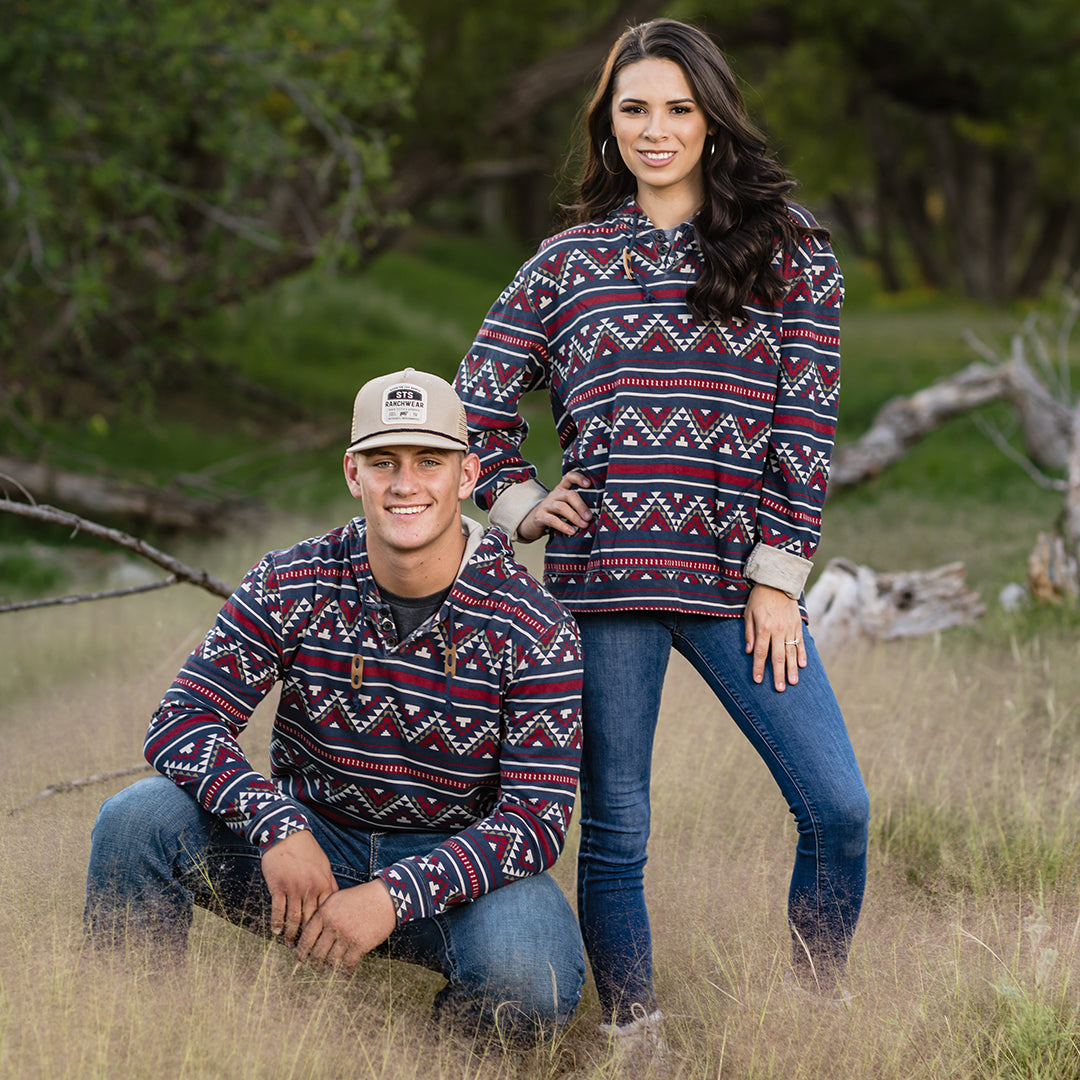Man kneeling next to woman in a field with both wearing blue jeans and a navy unisex hoodie with Aztec pattern featuring a drawstring hood, quarter button closure, and two exterior hand warming pockets.