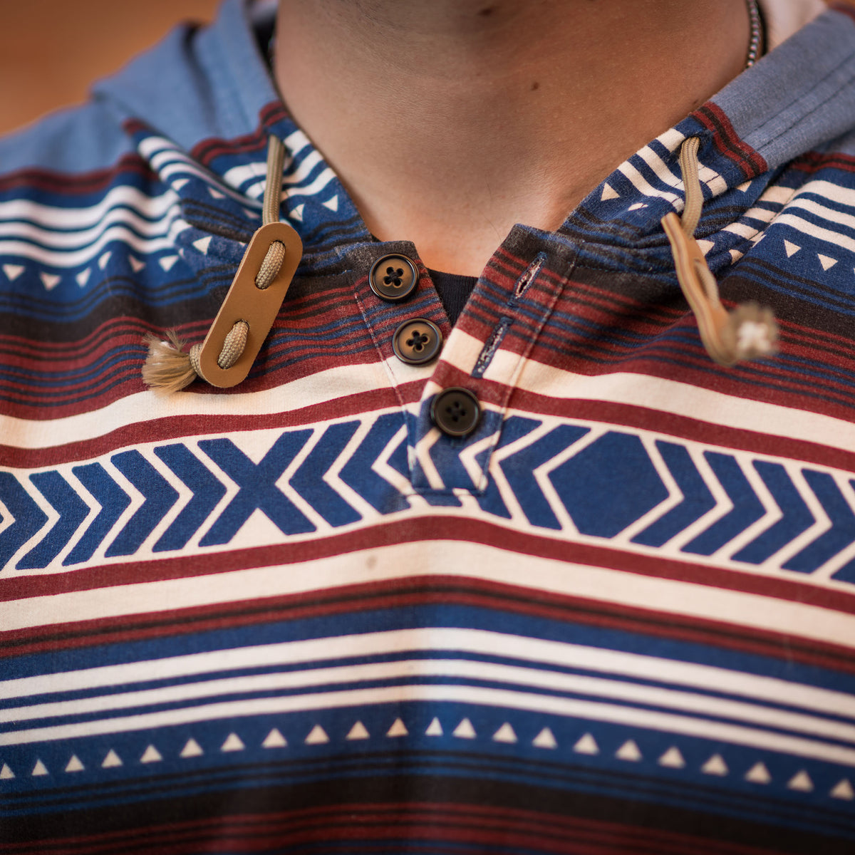 Detail of a unisex hooded sweatshirt with a geometric Aztec pattern in red, blue, and beige colors, featuring a quarter button closure and drawstring hood with leather pull tabs.