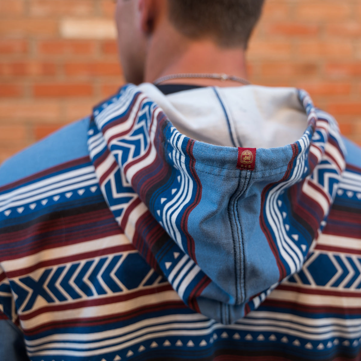 Detail of a unisex hooded sweatshirt with a geometric Aztec pattern in red, blue, and beige colors, featuring a drawstring hood.