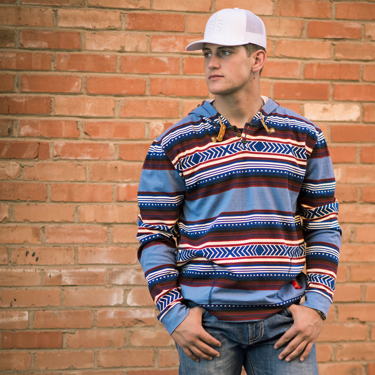 Person stand in front of a brick wall wearing a white hat, jeans and a unisex hooded sweatshirt with a geometric Aztec pattern in red, blue, and beige colors.