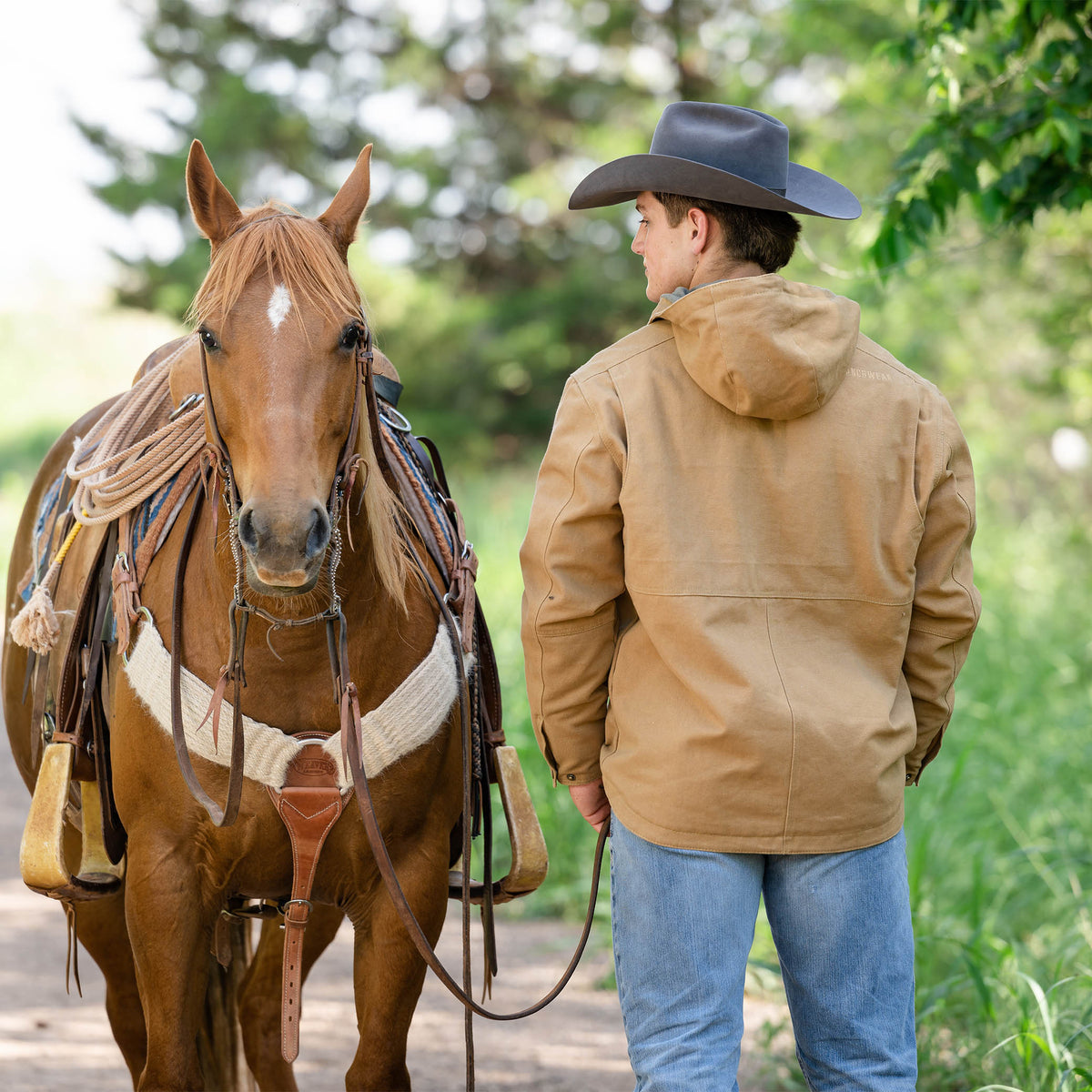 Man facing away wearing a gray cowboy hat, jeans and a beige, hooded, fleece-lined men's canvas work jacket standing next to a brown horse.