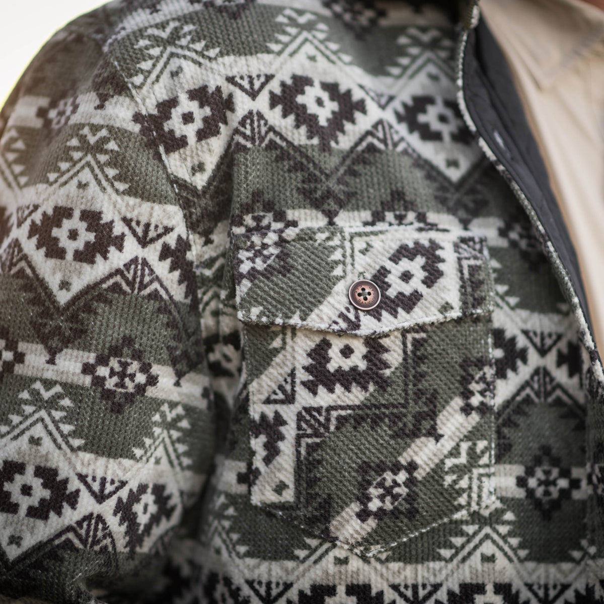 Detail of a men's reversible jacket with a geometric pattern in green, beige, and black colors, featuring button front closure and multiple pockets.